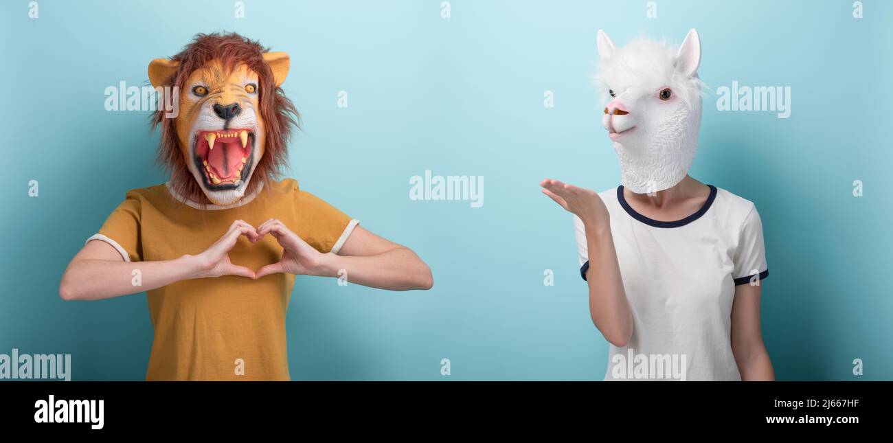 Young woman in lion mask shows love heart affection hand gesture, and woman in alpaca mask blows and send air kiss, sending love, isolated on blue bac Stock Photo