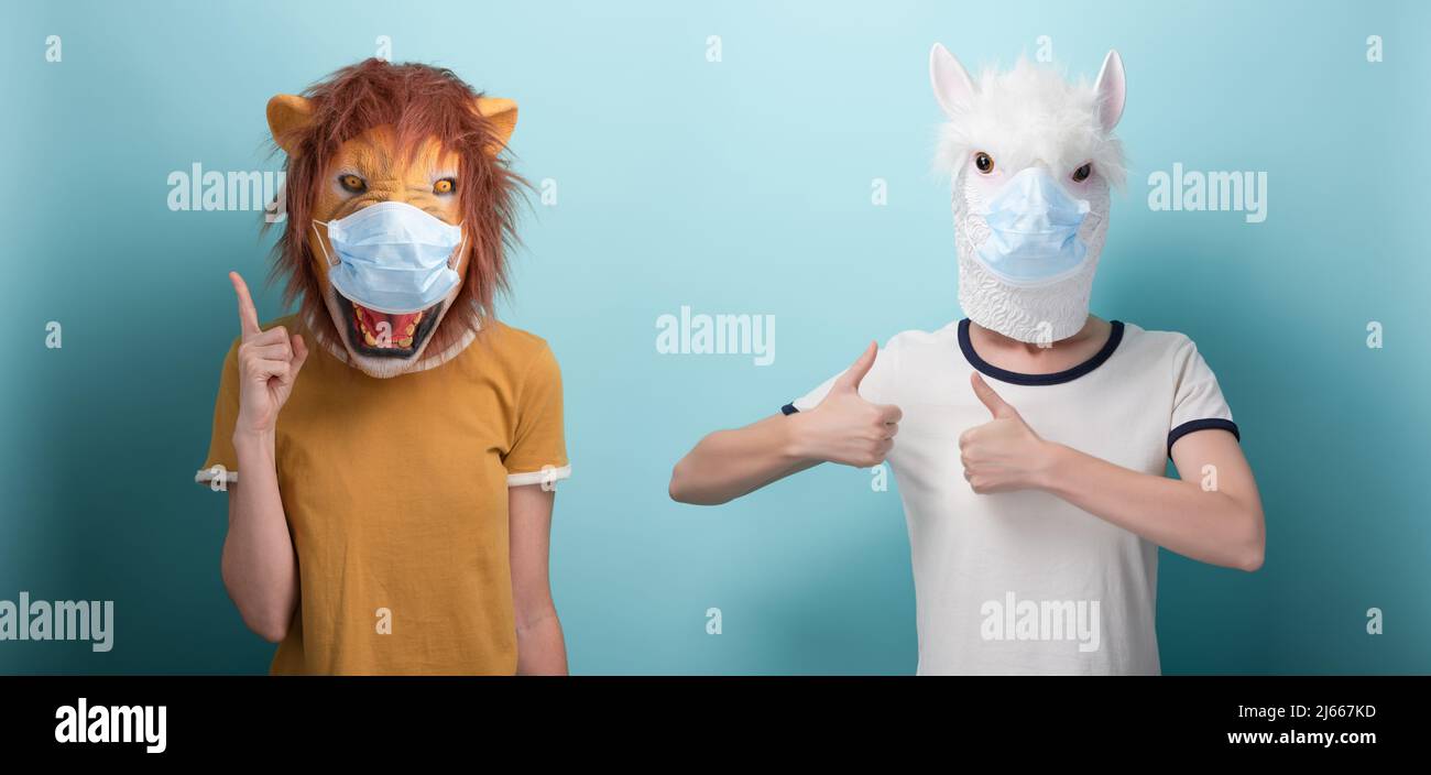 Young woman wearing lion and protection medical mask pointing up with index finger, and in alpaca mask keeps thumb up approval gesture Stock Photo