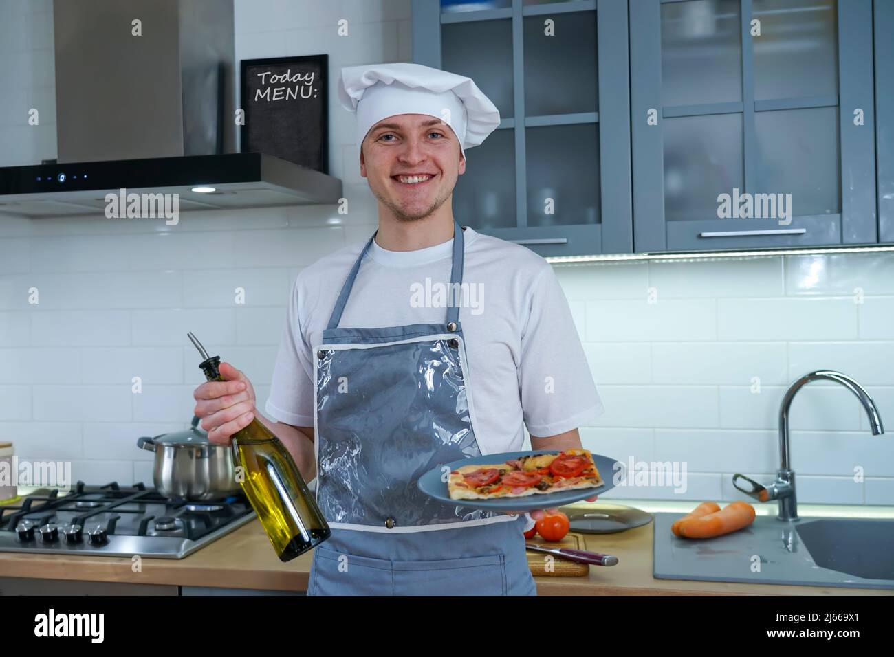 Portrait of a professional chef in the kitchen. He is holding a cooked pizza and a bottle of olive oil in his hands. Stock Photo