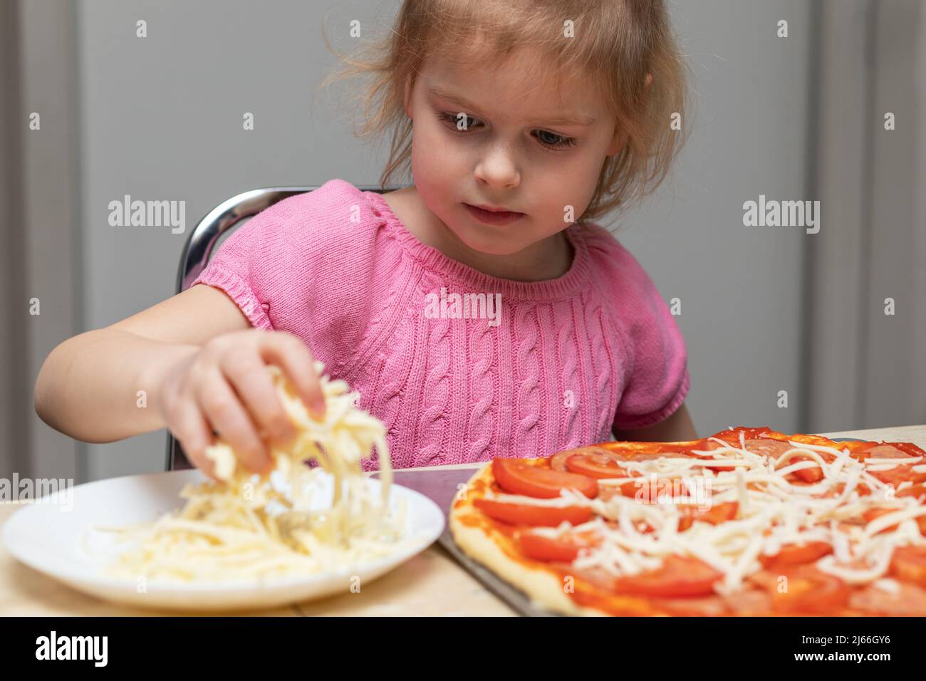 Small child cooking meal at home sitting at the table in the kitchen at home. Little girl making pizza, adding cheese. Stock Photo