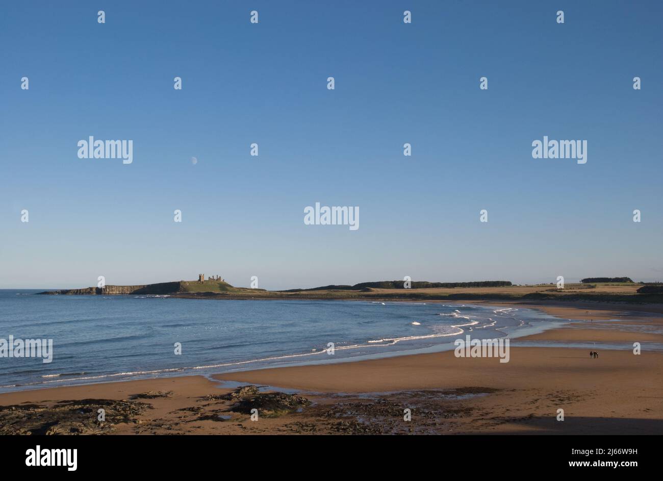Landscape image of the wide sweeping beach at Embleton leading to a distant Dunstanburgh Castle with moon above in a blue sky and few people Stock Photo