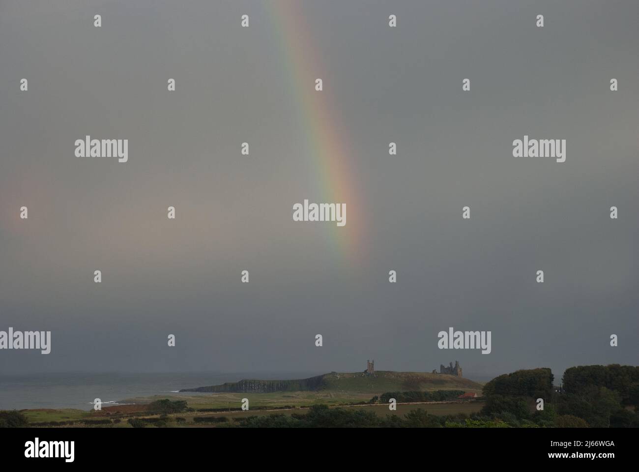 Landscape of Northumberland coast with  Dunstanburgh Castle in the far distance and rainbow arcing down in front of dark sky Stock Photo