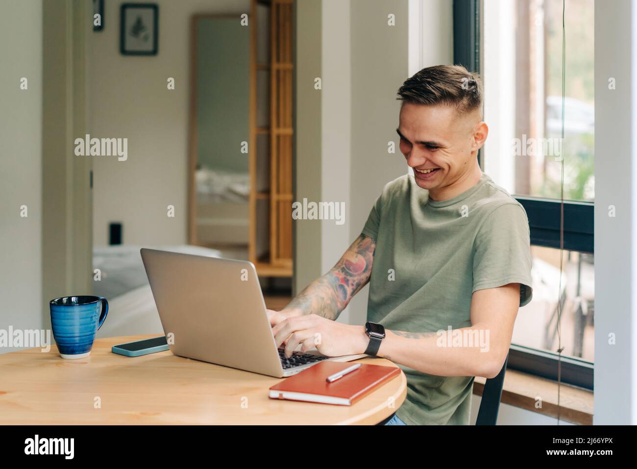 Cheerful laughing young man works on a laptop computer at a table at home. Stock Photo