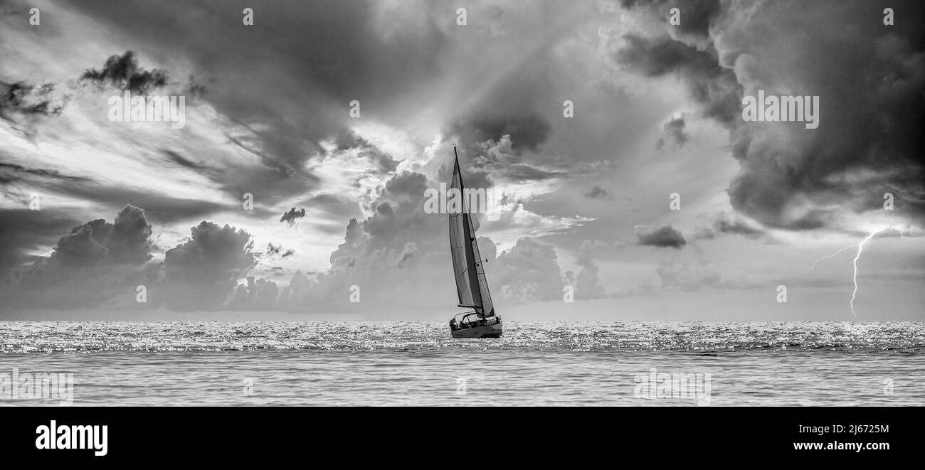 Sailboat navigating a silvery sea in a stormy cloudy sunset Stock Photo