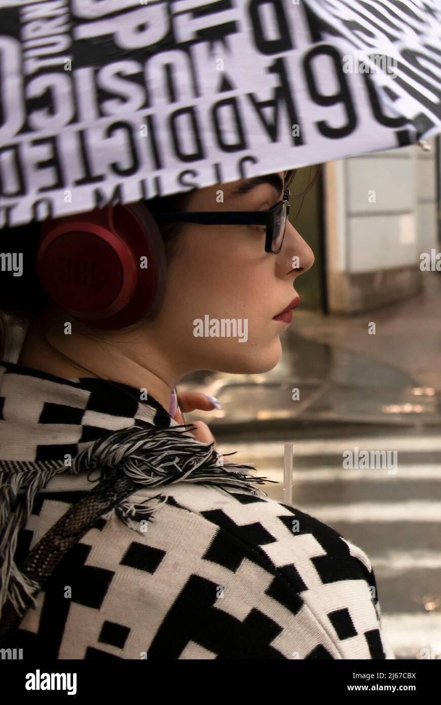 Belgrade, Serbia - April 27, 2022: Young woman in black and white pattern outfit wearing red headphones, red lipstick and eyeglasses under umbrella on Stock Photo