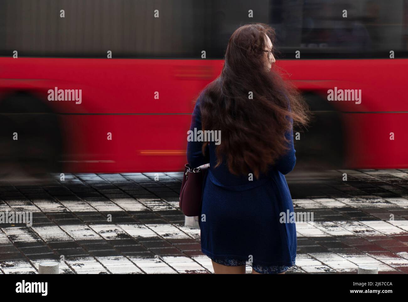 Belgrade, Serbia - April 27, 2022: Young woman with long wavy hair waiting on a crossroad on a rainy day with traffic in motion blur, rear view Stock Photo