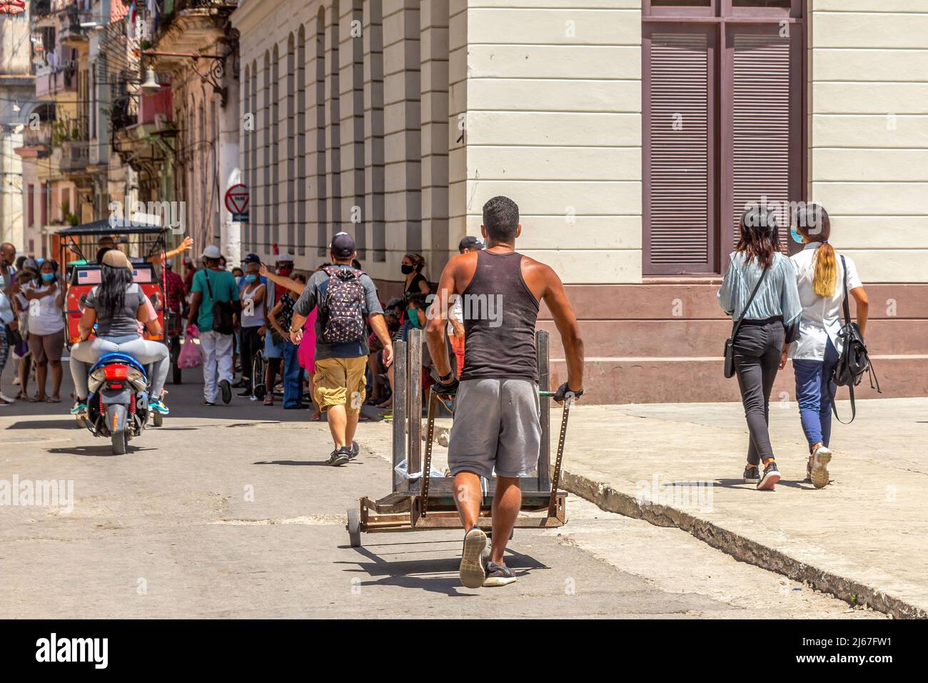 A Cuban man pushes a cart where carries an upside-down table. A large group of people is seen in the background. Stock Photo