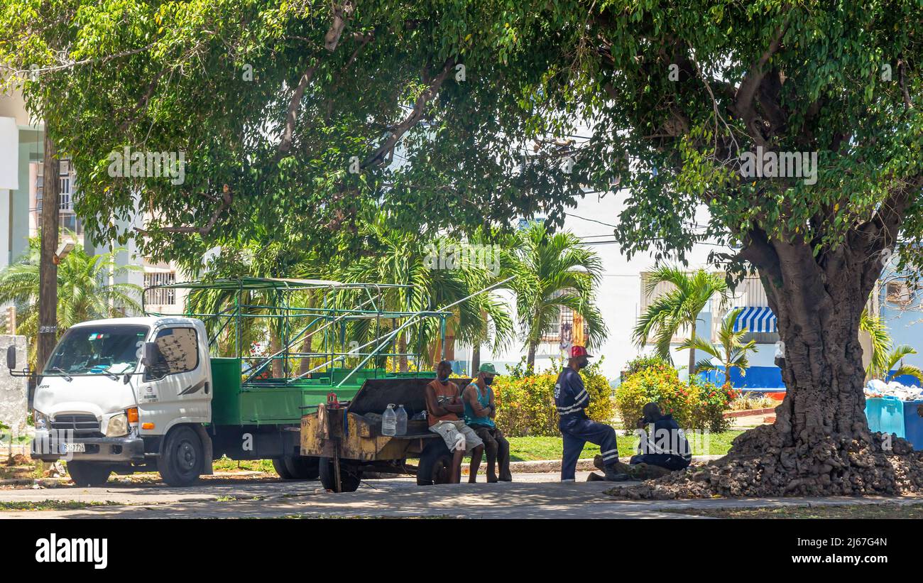 Construction workers take a break in the shade of a large tree. Their truck is parked across the street. Two of them wear uniforms reading Cupet. Stock Photo