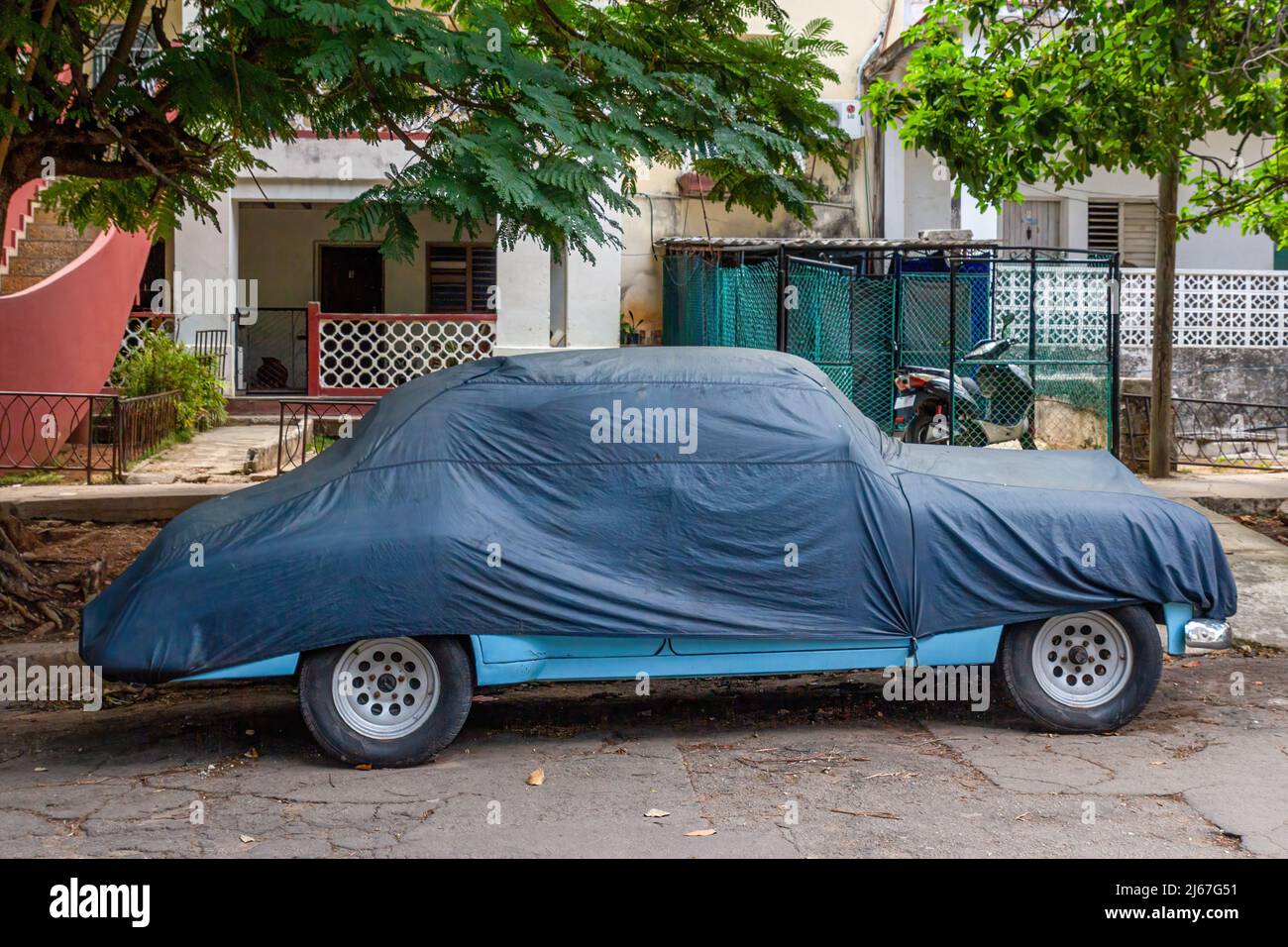 An old vintage American car is covered with a tarp while parking under a tree. Private houses and an electric bicycle are seen in the background. Stock Photo