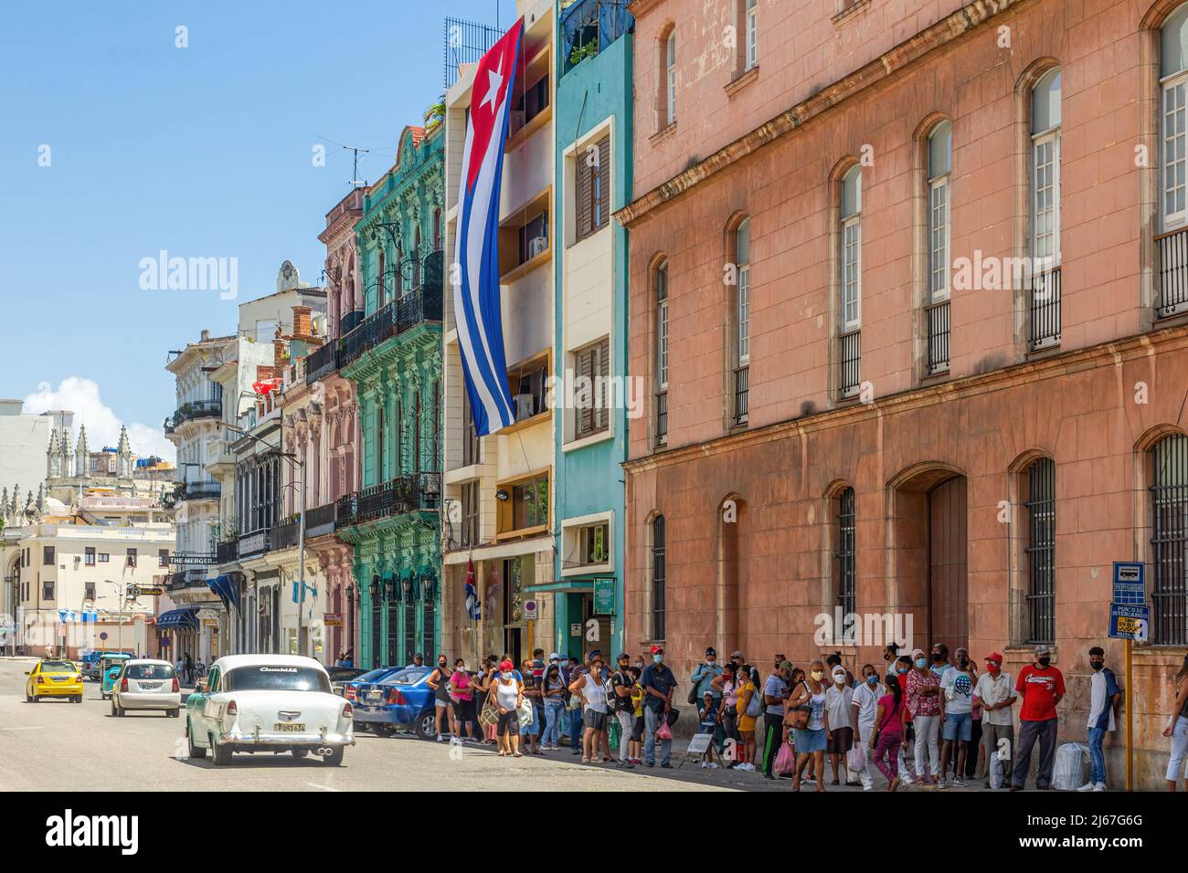 Large group of people waiting for a public omnibus in a bus stop. A large Cuban flag hangs from a government building. An old American car drive in th Stock Photo