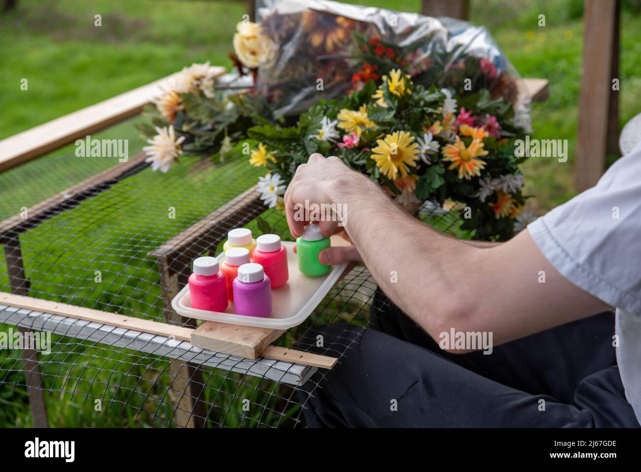 Adult arts and crafts art therapy creativity concept, man paints silk flowers in calm outdoor setting with green grass background and crutches leaning Stock Photo