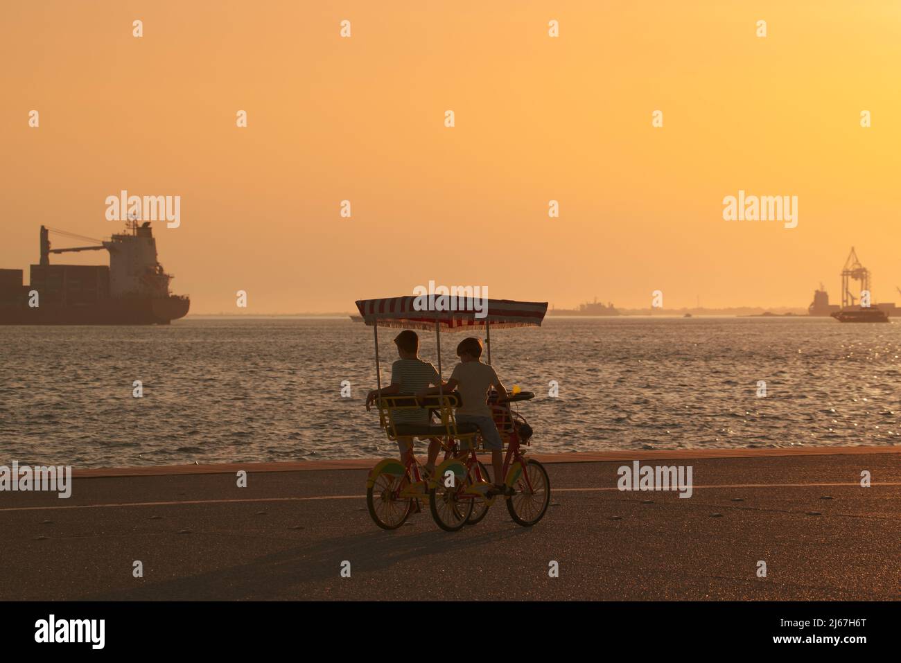 Two boys in a four-wheeled bicycle (surrey bike) at promenade watching a ship in the sea at sunset. Stock Photo