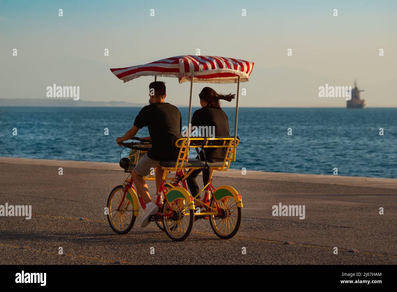 Young couple at sunset riding a four-wheeled bicycle (surrey bike) at promenade close to the sea. Stock Photo