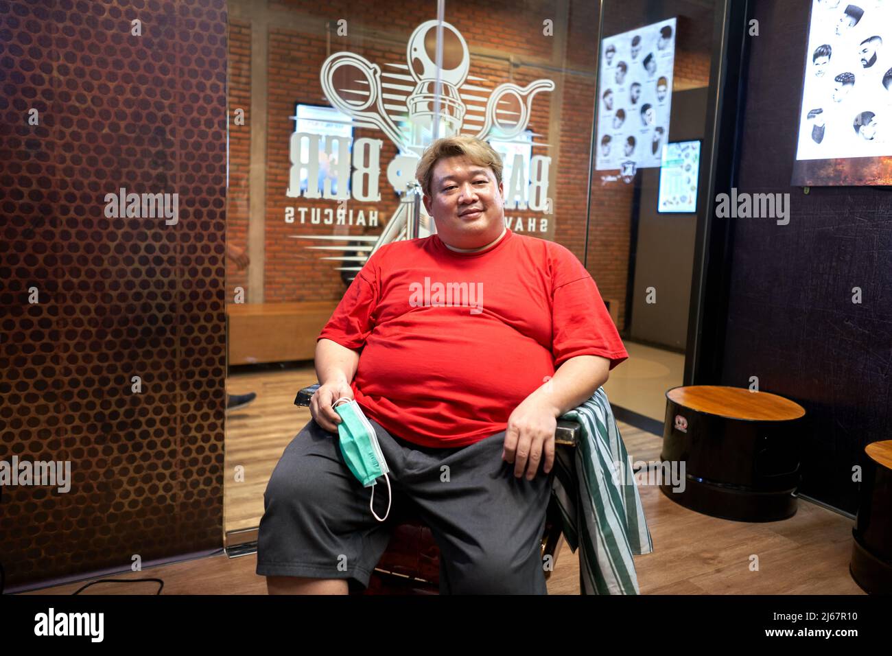 Portrait of a smiling asian obese barber sitting on a chair in a barber shop Stock Photo