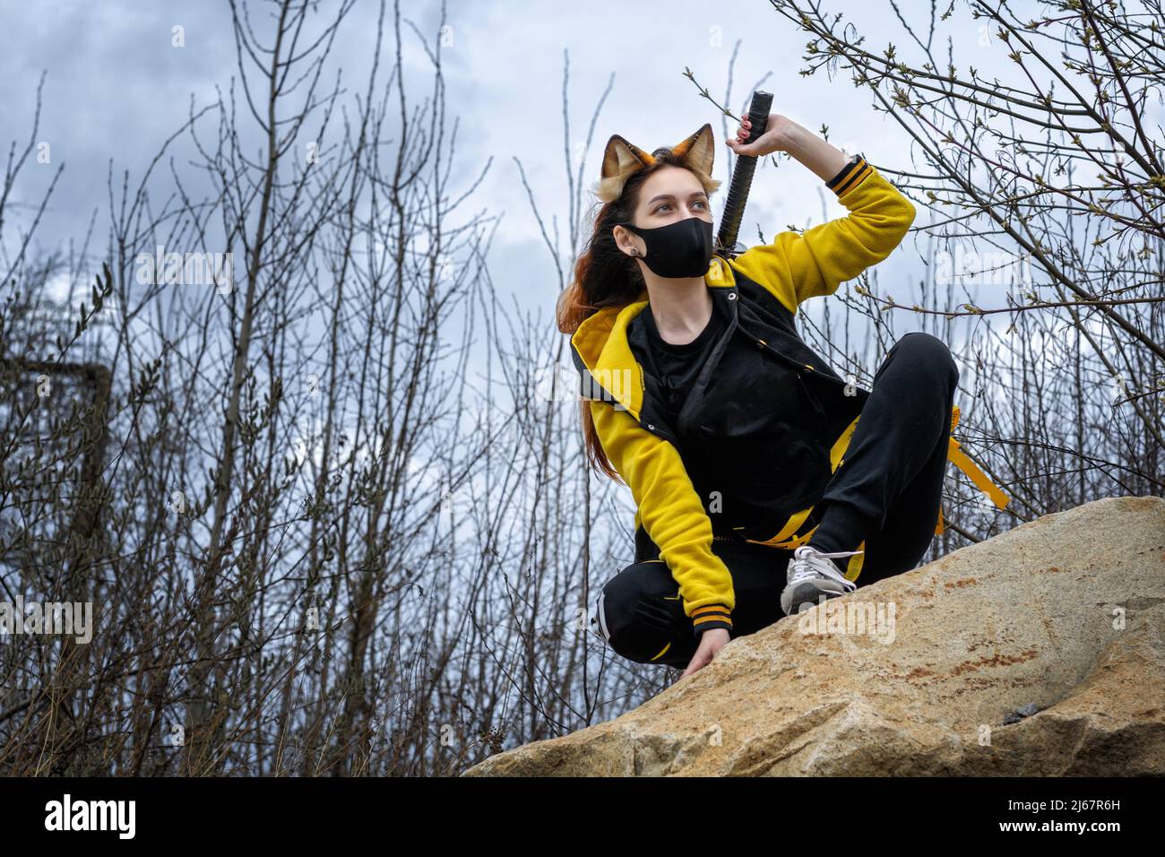 a girl in a yellow suit, mask and fox ears is sitting on a rock in the forest with a samurai sword in her hand Stock Photo