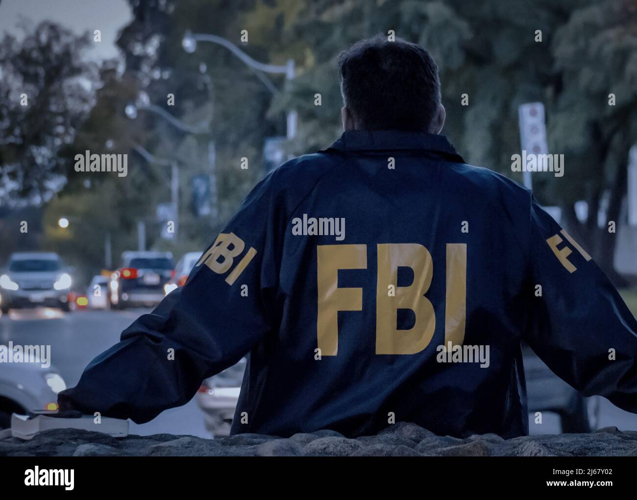 Male FBI agent wearing dark blue coat with FBI logo looking down the street with cars in the dusk seen from behind. Stock Photo