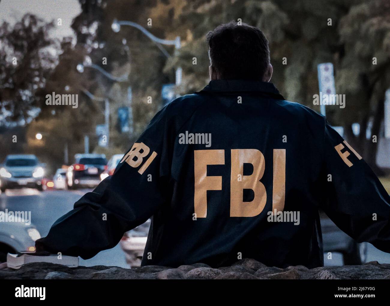 Male FBI agent wearing dark blue coat with FBI logo looking down the street with cars in the dusk seen from behind. Stock Photo