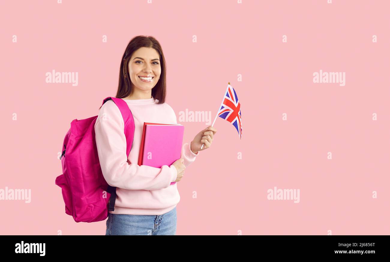Happy woman who's learning English standing on pink background, holding British flag and smiling Stock Photo