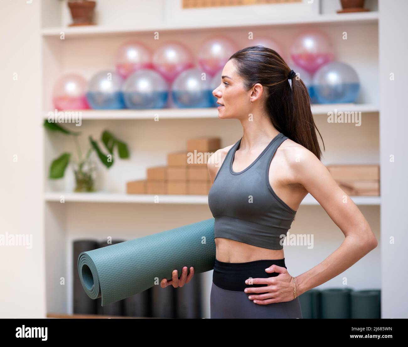 Slim female athlete with rolled up mat holding hand on waist and looking away at start of fitness workout in gym Stock Photo