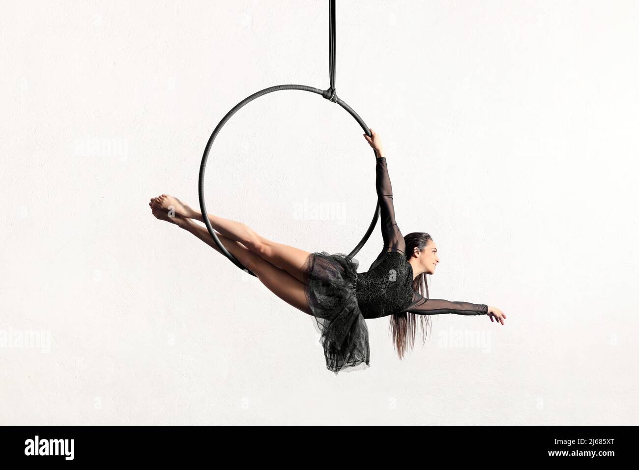 Full body of active barefoot female gymnast doing siren pose on hanging aerial hoop or lyra against white background in light studio Stock Photo