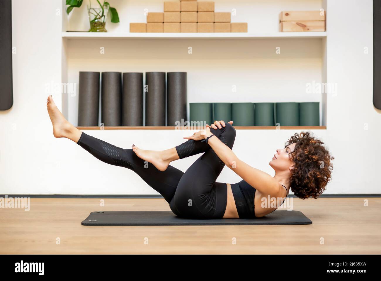 Side view of barefoot woman in sportswear with curly hair doing knee to chest exercise on floor during functional training in gym. Fitness concept. Stock Photo