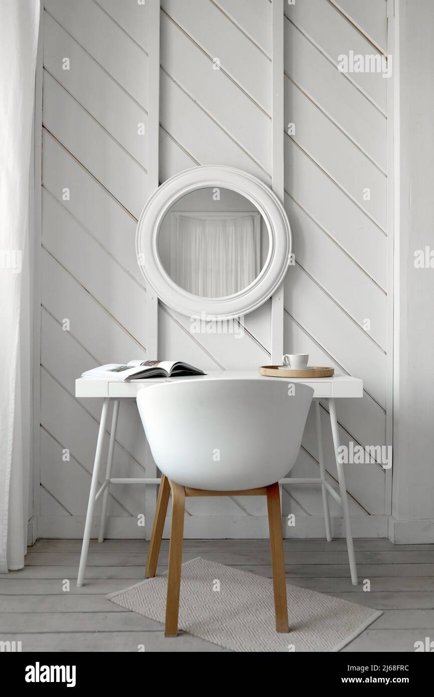 White chair at table with book and cup of beverage placed near wall with round mirror in light room designed in minimal style Stock Photo
