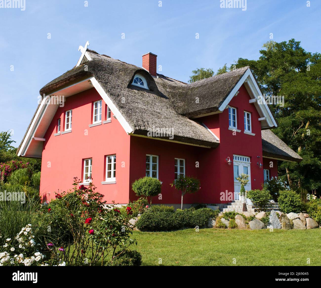 Red house with a thatched roof in beautiful nature. Green grass and trees in a property. Traditional real estate in Europe on a sunny summer day. Stock Photo