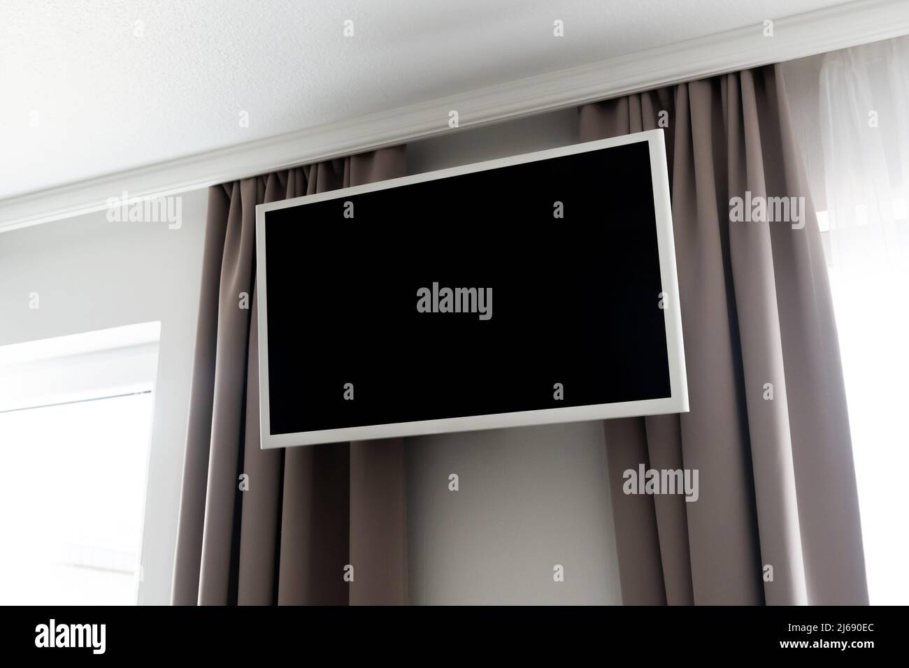 Empty TV screen in a hotel hanging on the wall. Bedroom television as design mockup template in a motel. Curtains and windows next to the display. Stock Photo