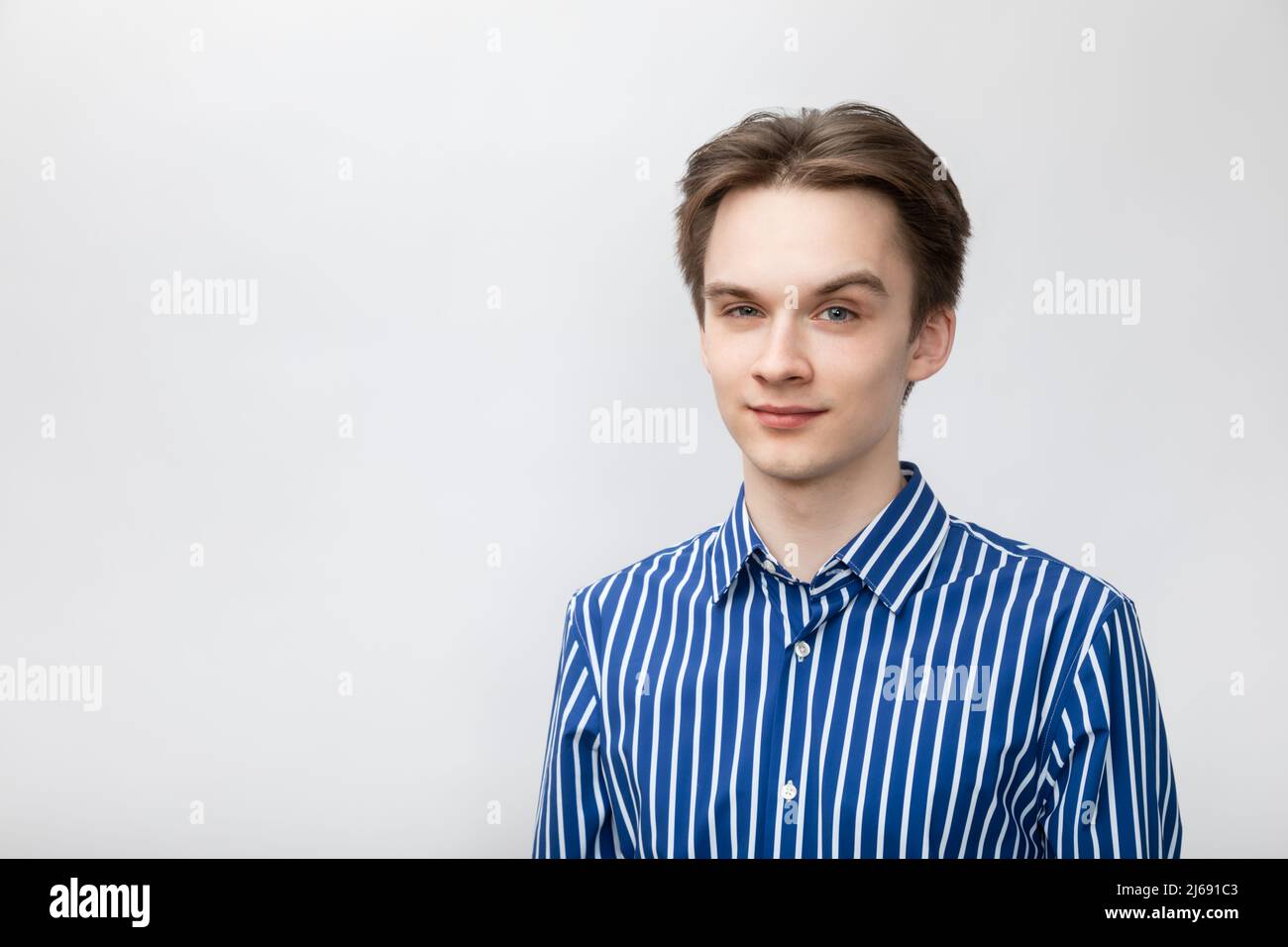 Portrait of confident and cheerful young man wearing blue-white striped button shirt looking at camera with one eyebrow raised. Studio shot on gray ba Stock Photo
