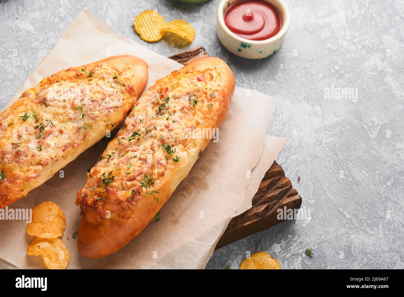 Baguette boats. Hot baked sandwich on baguette bread with ham, bacon, vegetables and cheese on parchment and wooden stand on gray concrete background. Stock Photo