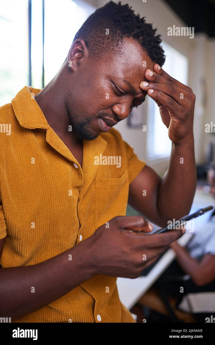 A Black man rubs his forehead looking confused while on his mobile device Stock Photo