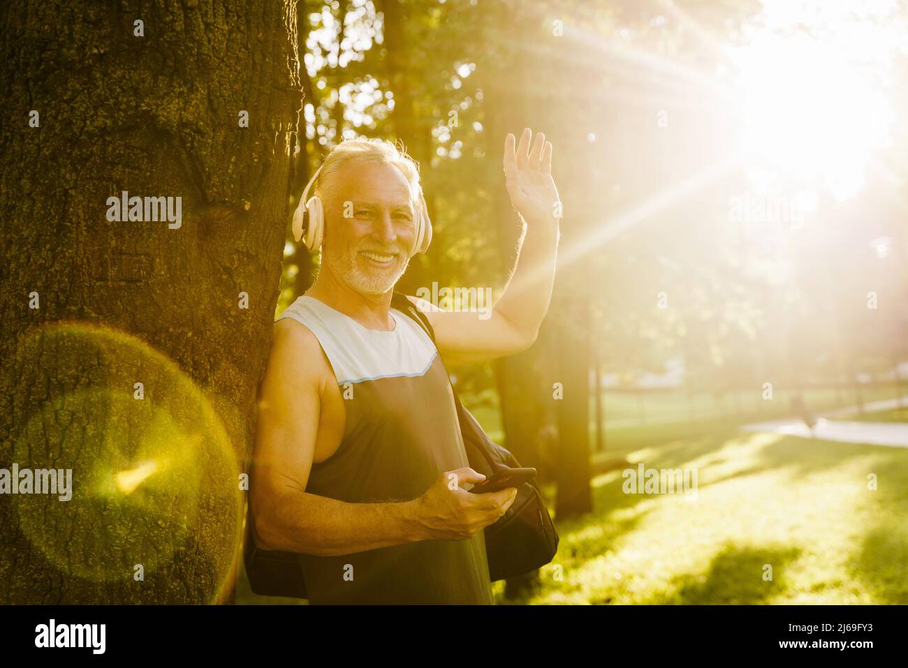 Grey senior man smiling and waving hand while using cellphone in park Stock Photo