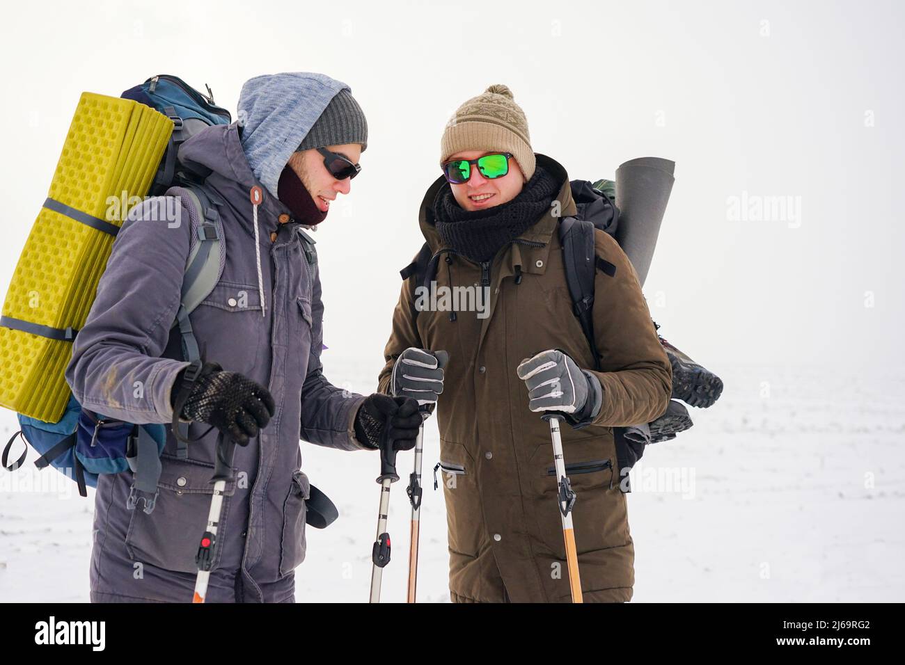 Two guys walk through loose snow during a winter expedition. They carry large backpacks, warm jackets. They hold trekking sticks in their hands. Stock Photo