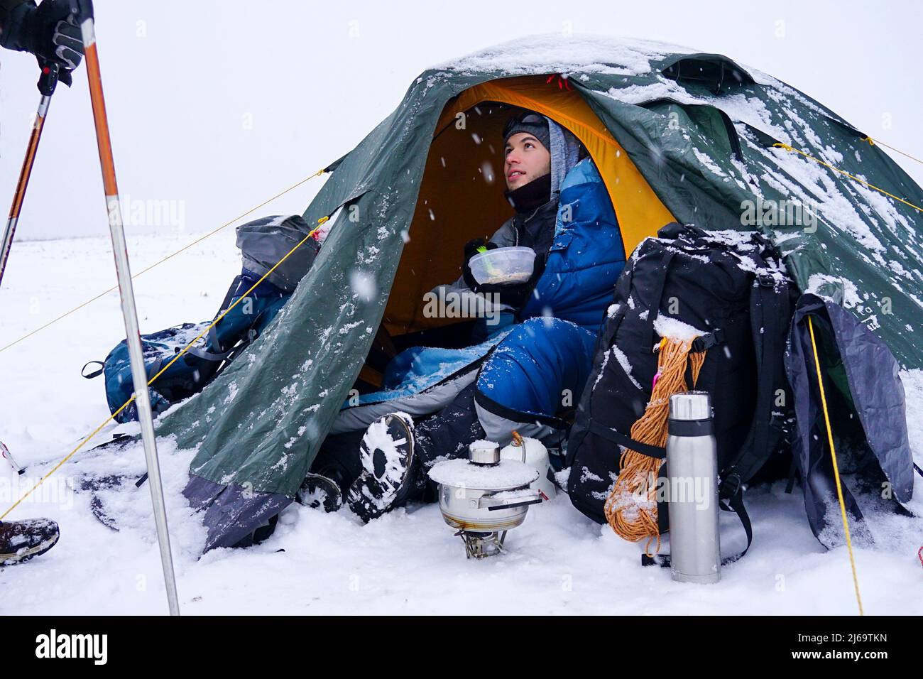 A tourist sits near a tent set on the snow, he takes out a Cord from a backpack for additional fixation of the tent during a winter storm Stock Photo