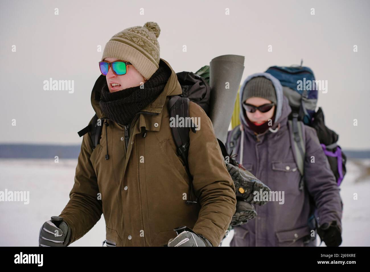 Two guys walk through loose snow during a winter expedition. They carry large backpacks, warm jackets. They hold trekking sticks in their hands. Stock Photo