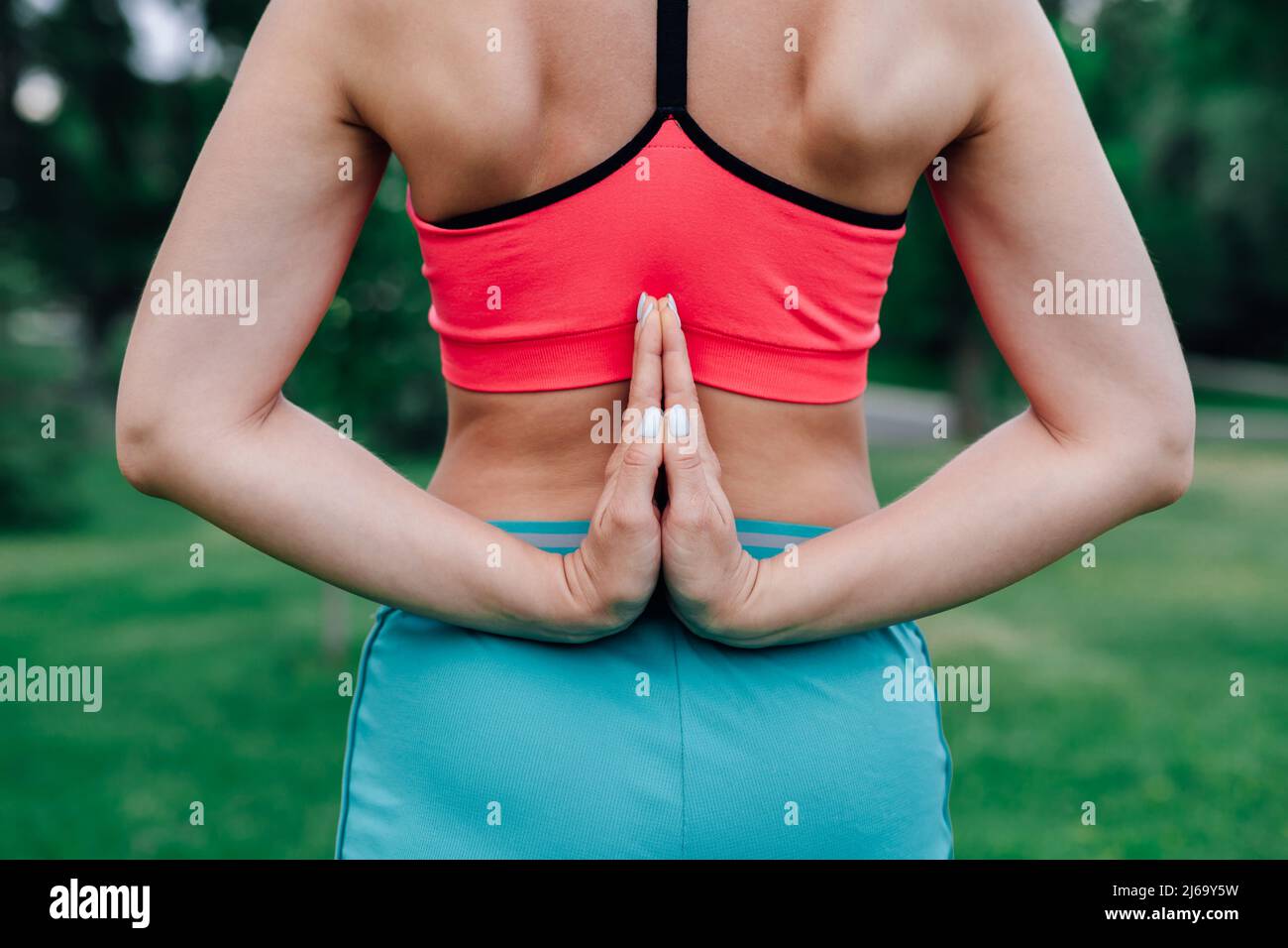 Exercises for proper posture. Body of young woman in pink top and blue sports trousers with her palms folded behind her back, standing in pose of Stock Photo