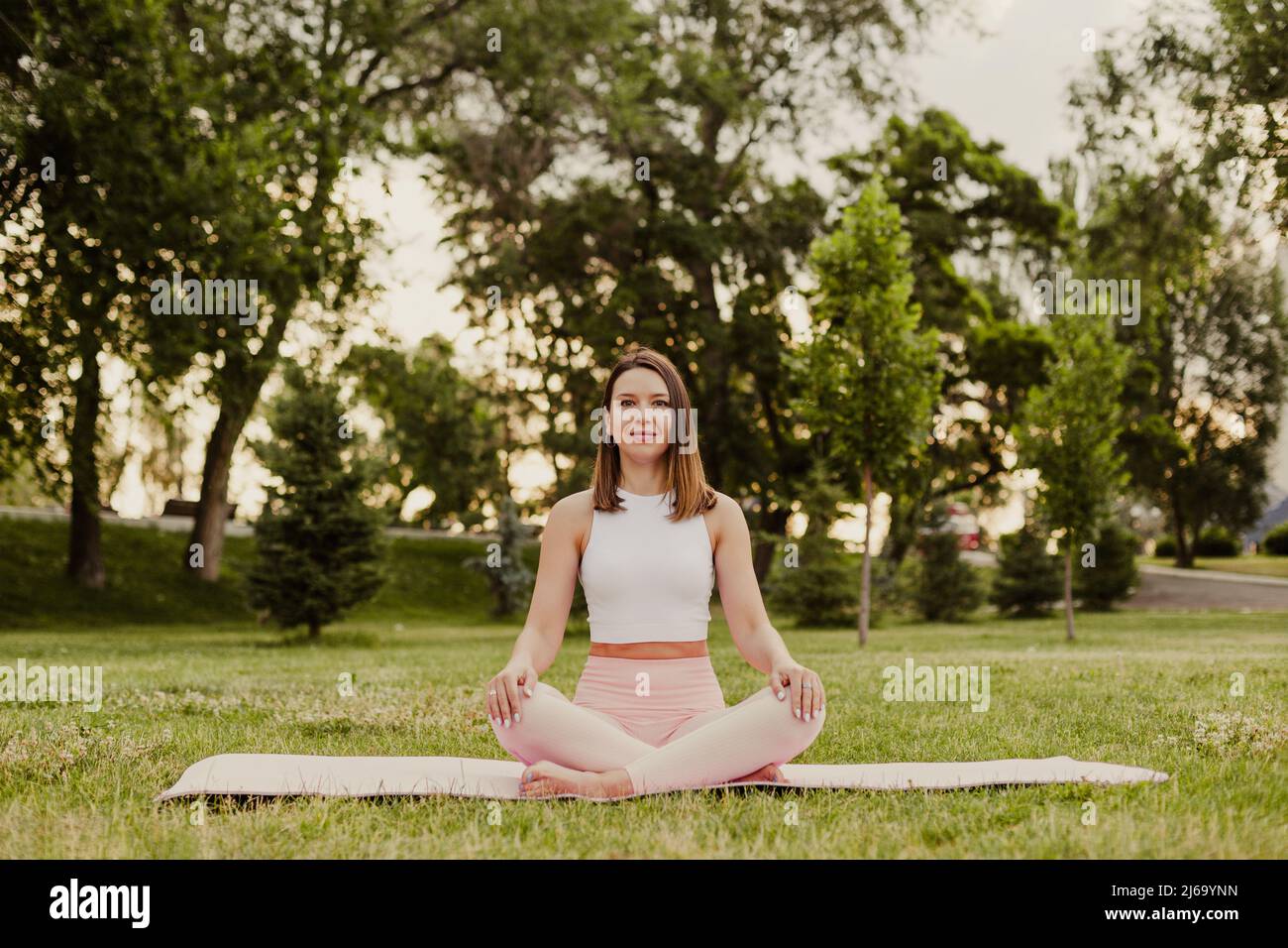 Young sport woman practice yoga on gymnastic mat in green park on grass. Sit cross legged in sportswear, do exercises to keep fit in summer morning Stock Photo