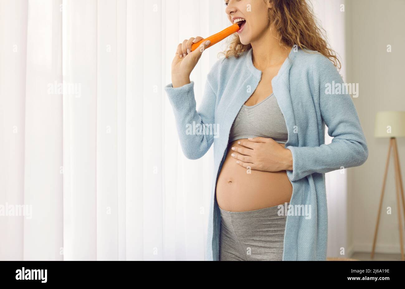 Happy beautiful young pregnant lady standing by the window at home and eating a carrot Stock Photo