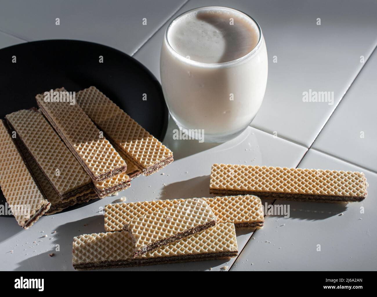 wafer cookies on a black plate and on a white table, with a glass of cold milk on the side lit by the sunlight coming from a window Stock Photo