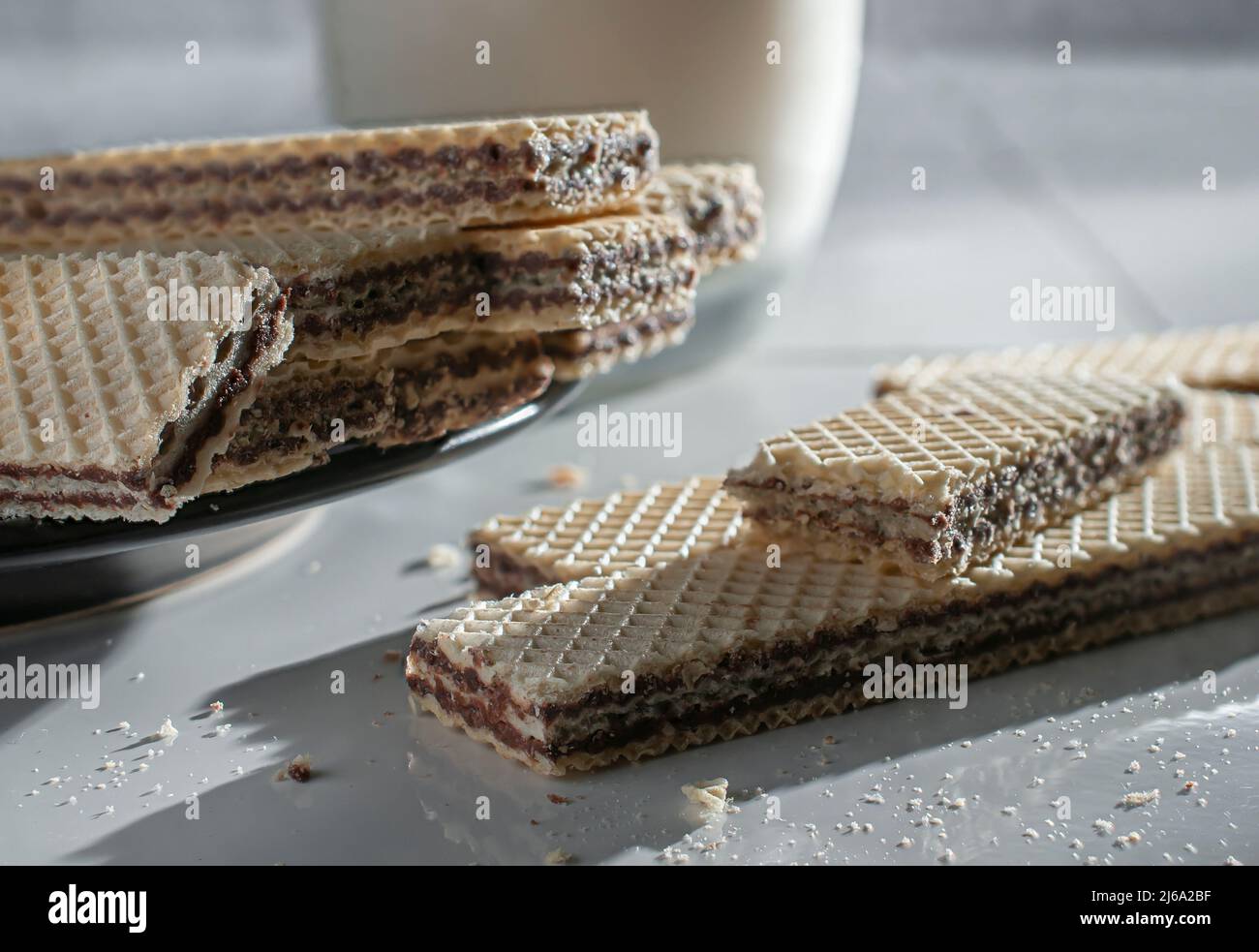 wafer cookies on a black plate and on a white table, with a glass of cold milk on the side lit by the sunlight coming from a window Stock Photo