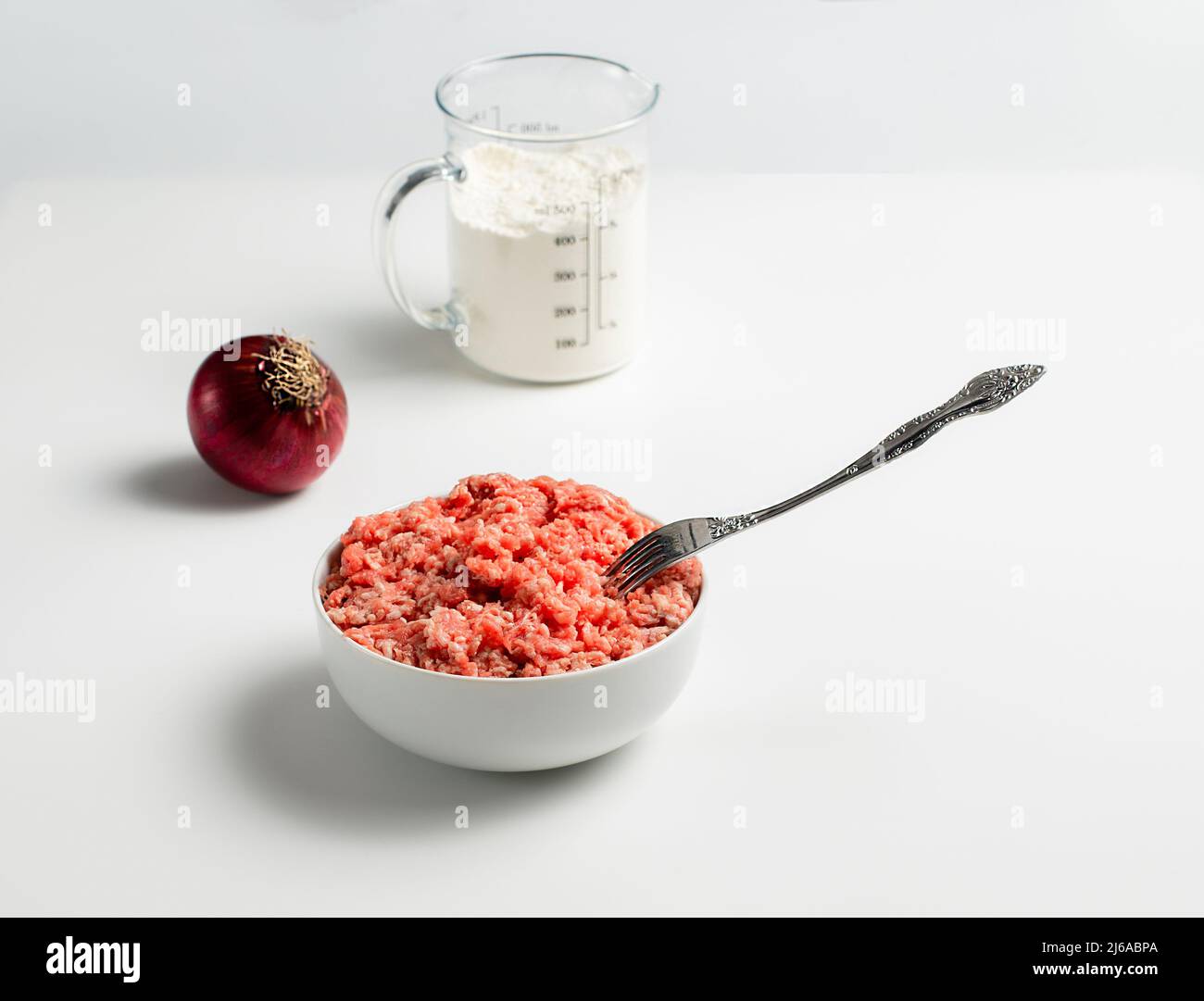 Plate with fresh ground beef and fork, red onion, measuring cup with flour on a white background, space for text, stock photo Stock Photo