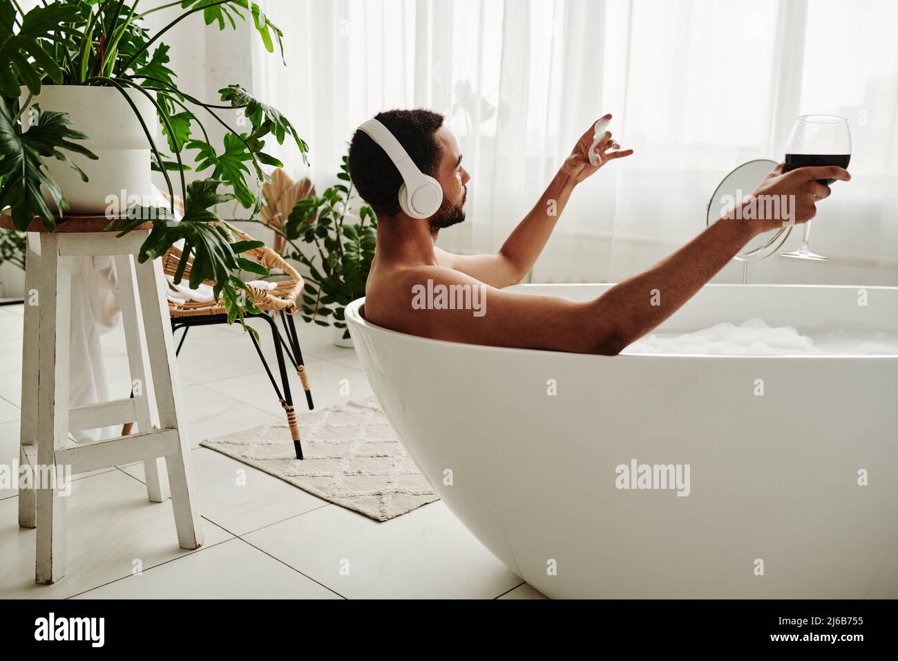 Young man relaxing in bathtub with glass of red wine and enjoying music