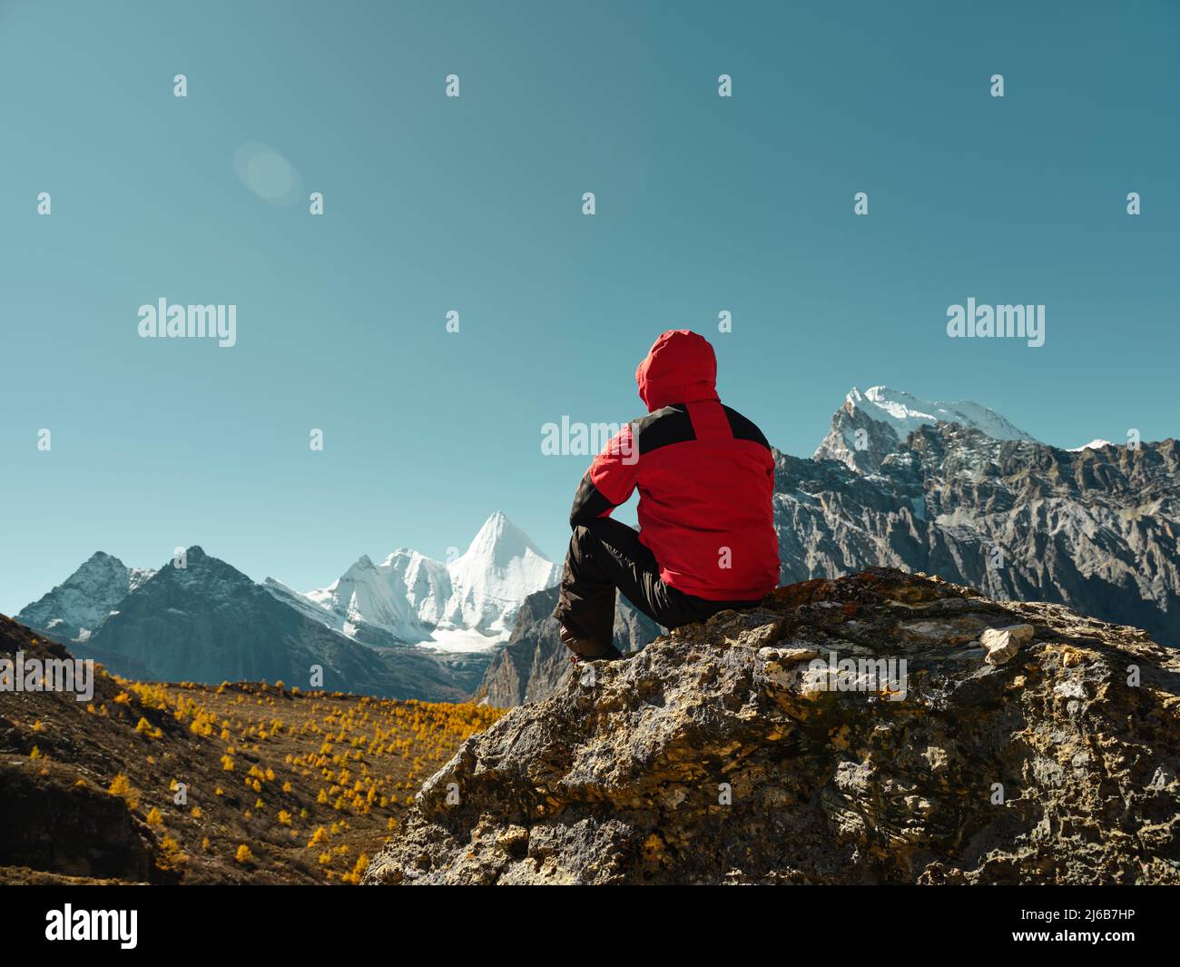 rear view of asian man sitting on top of rock with Yangmaiyong (or Jampayang in Tibetan) mountain peak in the distance in Yading, Daocheng County, Sic Stock Photo