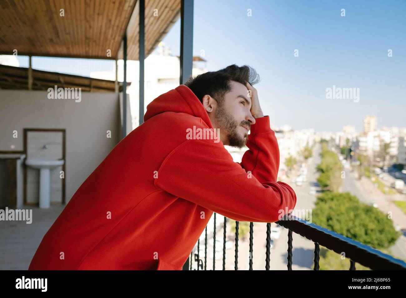 Alone stressed man wearing red hoodie on balcony. Serious man thinking about something. Unpleasant pain. Sad unhappy handsome man holding his forehead Stock Photo