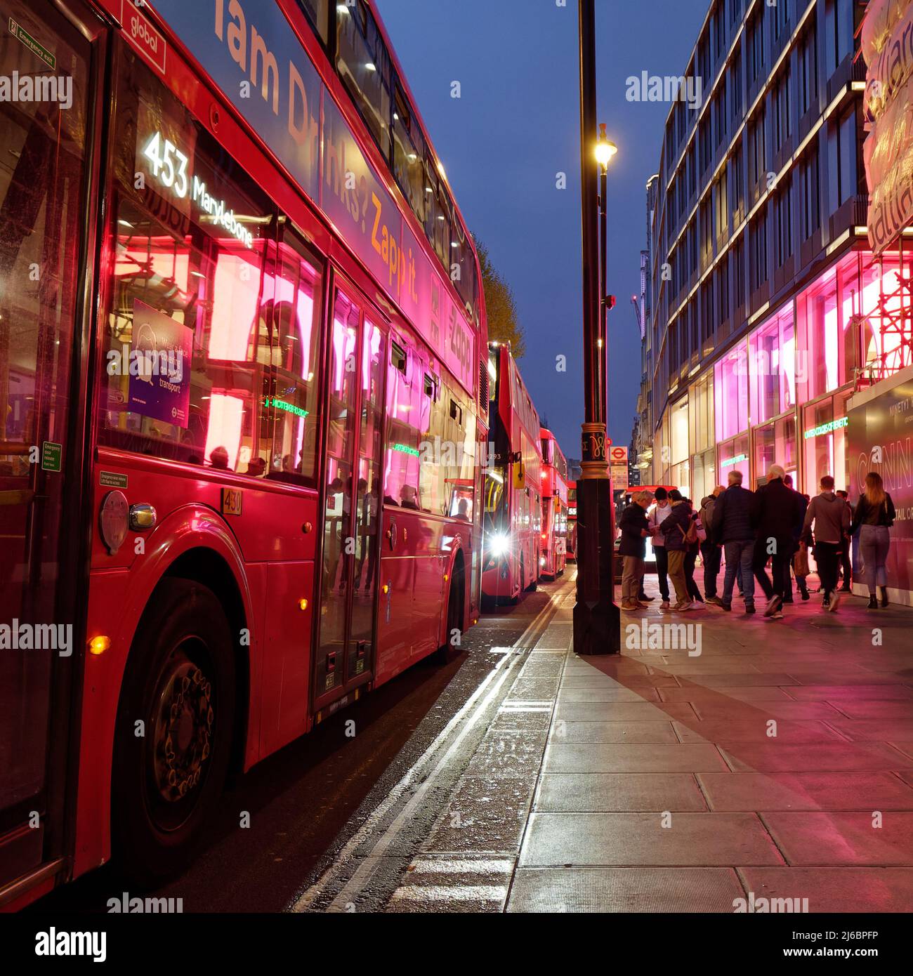 London, Greater London, England, April 23 2022: Street and property lights reflect onto a bus on Oxford Street as pedestrians pass by. Stock Photo
