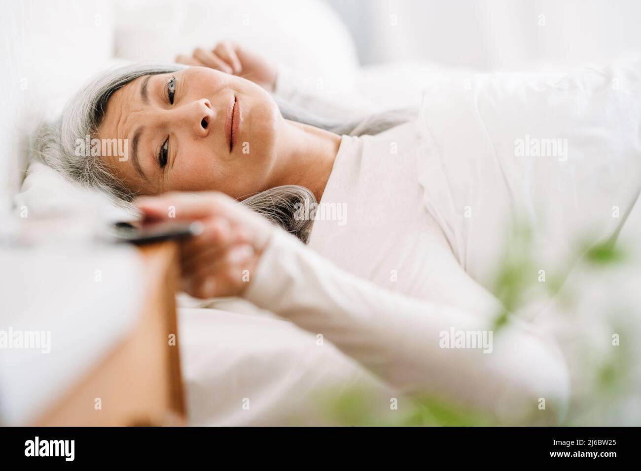 Grey asian woman using mobile phone while lying in bed after sleep at ...