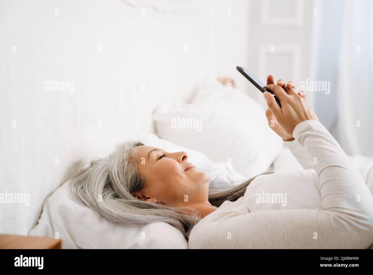 Grey asian woman using mobile phone while lying in bed after sleep at ...