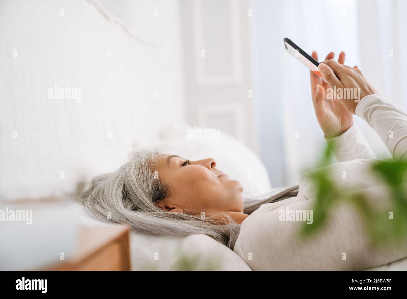 Grey asian woman using mobile phone while lying in bed after sleep at ...