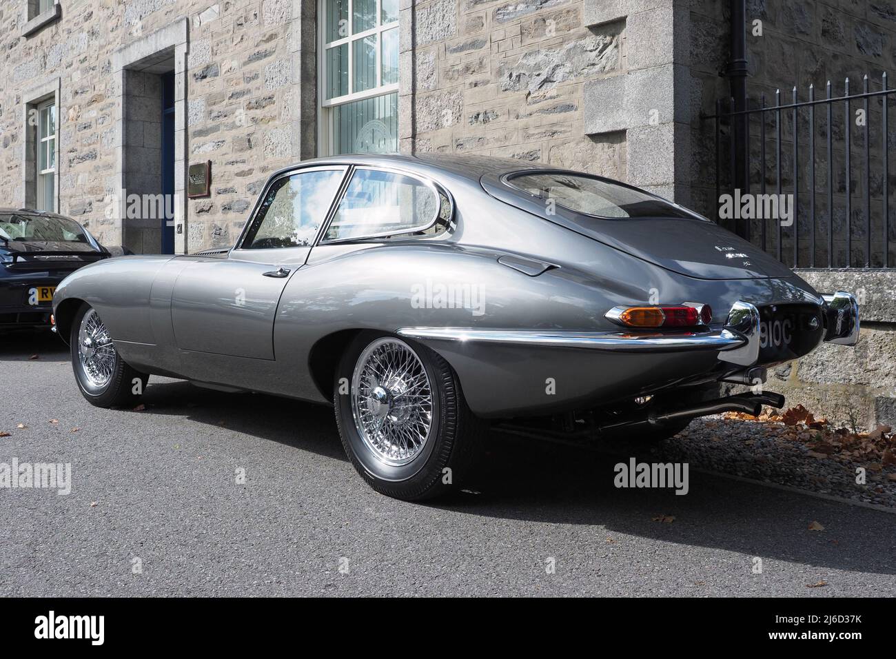 Rear view of a Jaguar E-Type, Series 1 coupe, with a straight six 4.2lt engine, in metallic pearl grey silver. Parked next to old stone building. Stock Photo