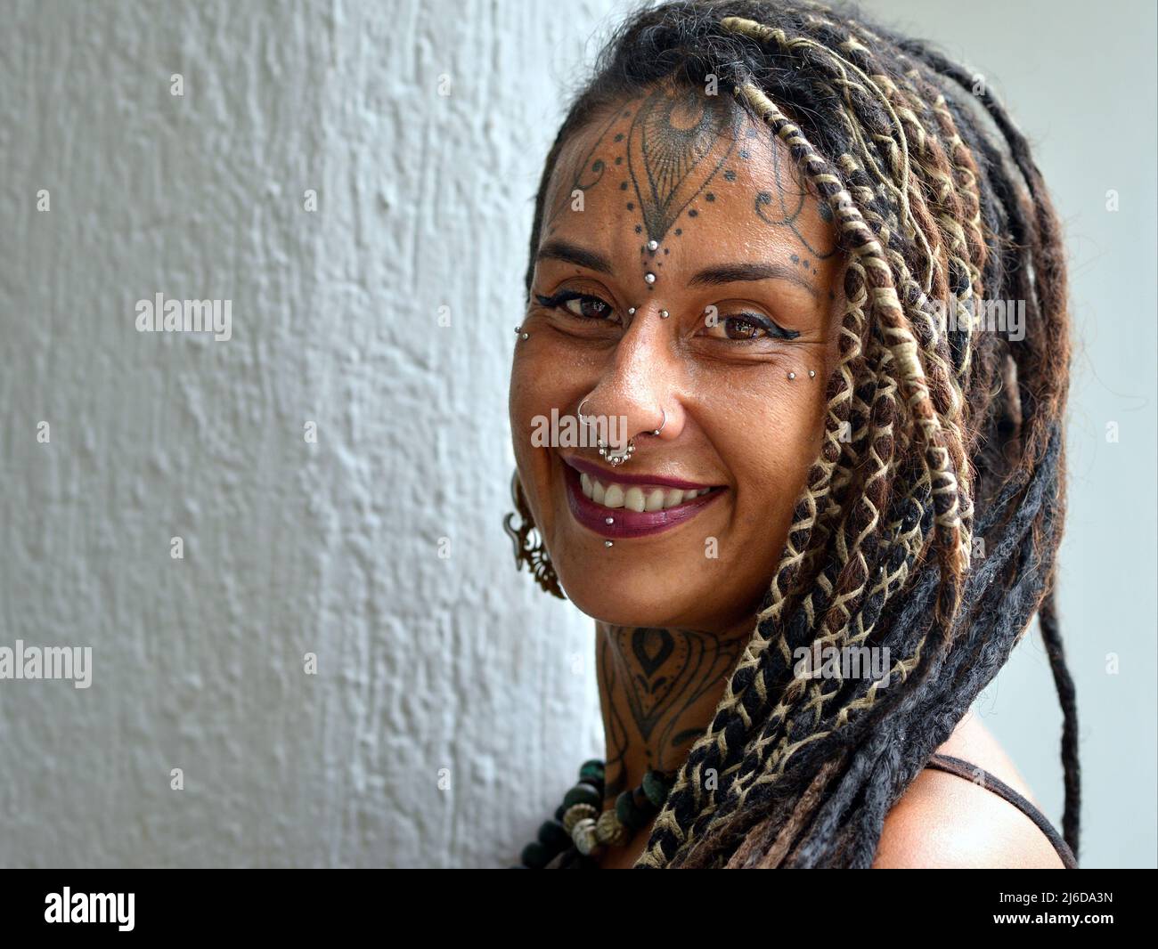 Attractive alternative young Mexican woman with long dread Rasta braids, body art facial piercing and tattoo smiles positively optimistic at viewer. Stock Photo